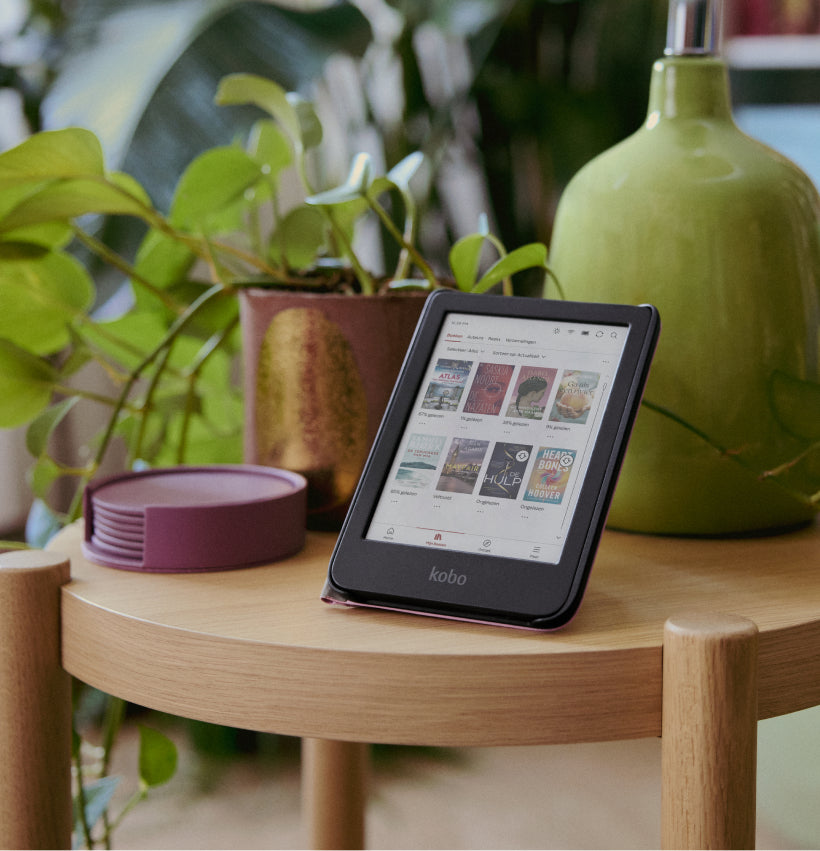 Een Kobo Clara Colour eReader in zijn SleepCover standaard, op een houten tafel bij een plant en een lamp.