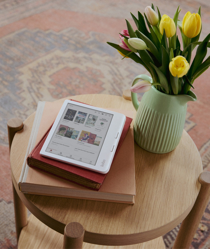 Een Kobo Libra Colour eReader op twee hardcover boeken, op een houten tafel met bloemen in een groene vaas.