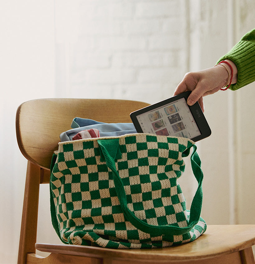 Een hand haalt een Kobo Clara Colour eReader uit een boekentas die op een houten stoel staat.