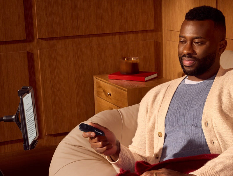 Een man, comfortabel zittend, houdt de Kobo Remote in zijn rechterhand, terwijl hij naar een Kobo eReader op een buigbare standaard kijkt.