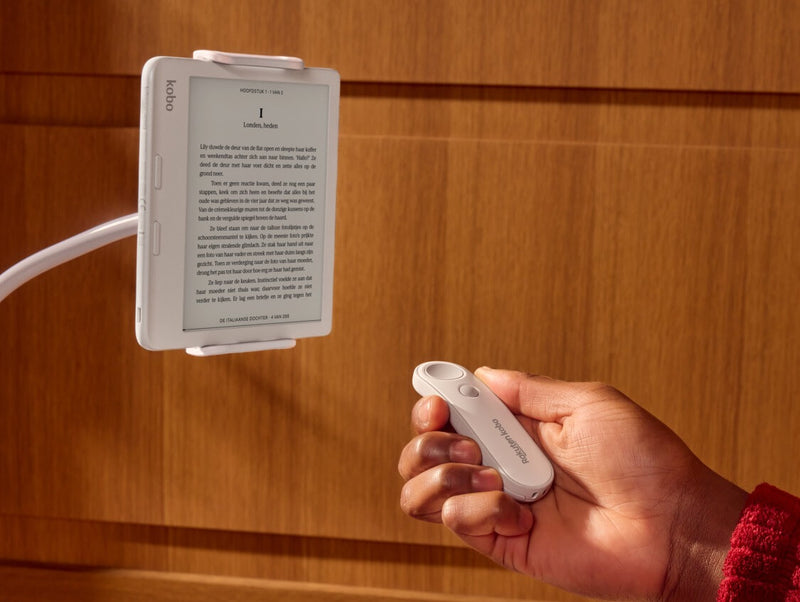 Een witte Kobo Remote wordt getoond in iemands rechterhand, wijzend naar een witte Kobo Libra Colour eReader op een buigbare standaard.